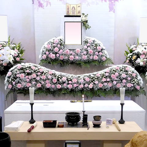 白やピンク、紫などたくさんの花で作られた祭壇と、その横にはスタンドの花が置かれ、手前には棺が置かれています