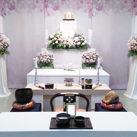 白やピンクの花で作られた祭壇と、その横にはスタンドの花が置かれ、手前には棺が置かれています